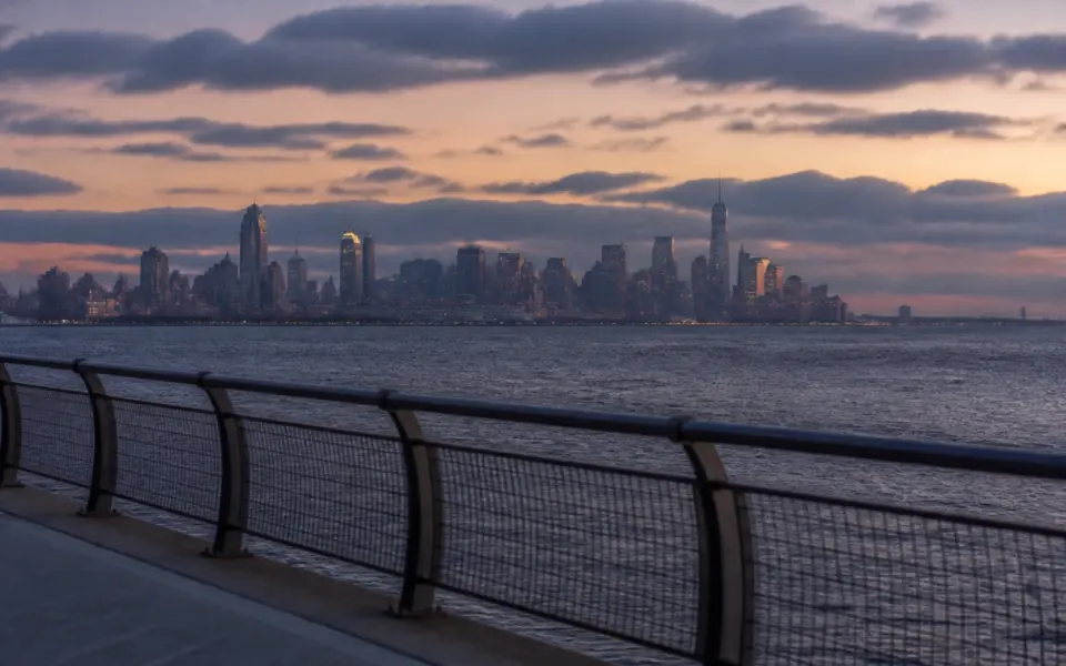 Illustration: the skyline of a distant city across open water at dusk, seen from a boardwalk railing