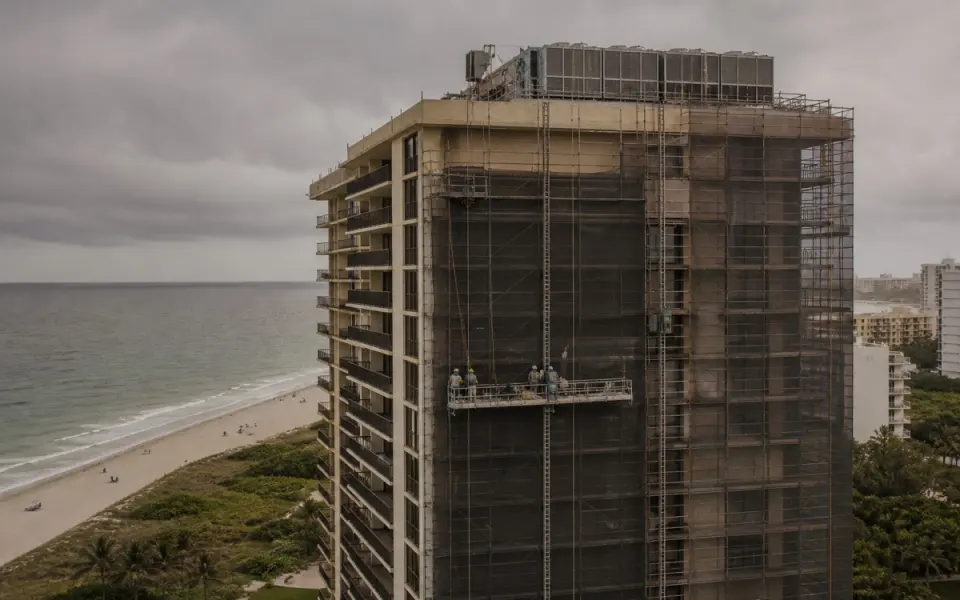 Illustration: a beachfront hotel tower wrapped in restoration scaffolding under an overcast sky